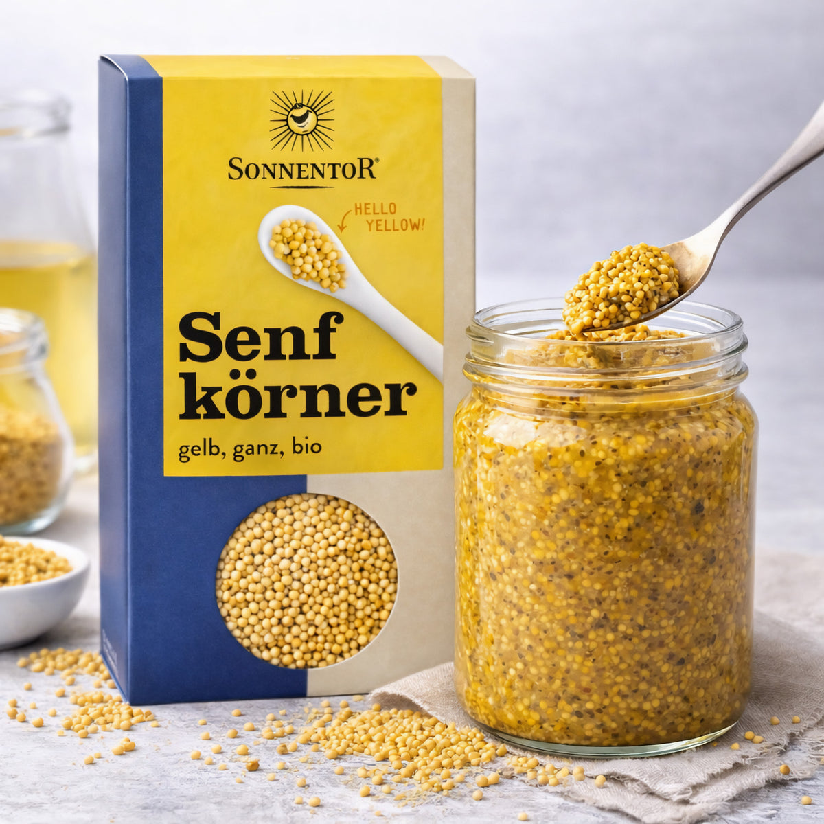 Sonnentor Senfkörner gelb ganz neben einem Glas mit angerührtem Senf, ein Löffel mit Senfmasse wird herausgehoben.