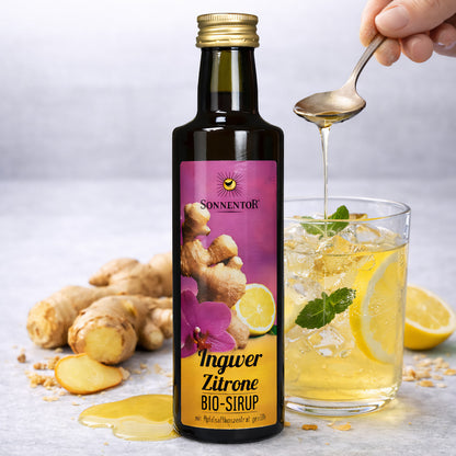 Getränkesirup von Sonnentor vor Glas mit Eis, Zitronenscheiben und Minze, daneben Ingwer und Zitrone, Sirup wird mit Löffel ins Glas gegeben.