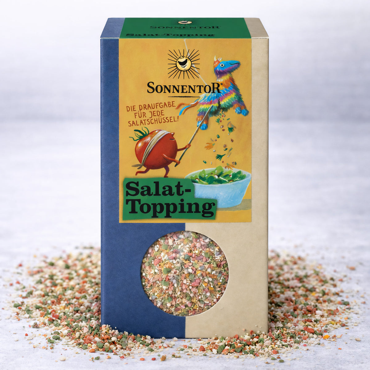 Packung Sonnentor Gewürzzubereitung Salattopping mit Sichtfenster, umgeben von verstreuten Gewürzen auf hellem Untergrund.