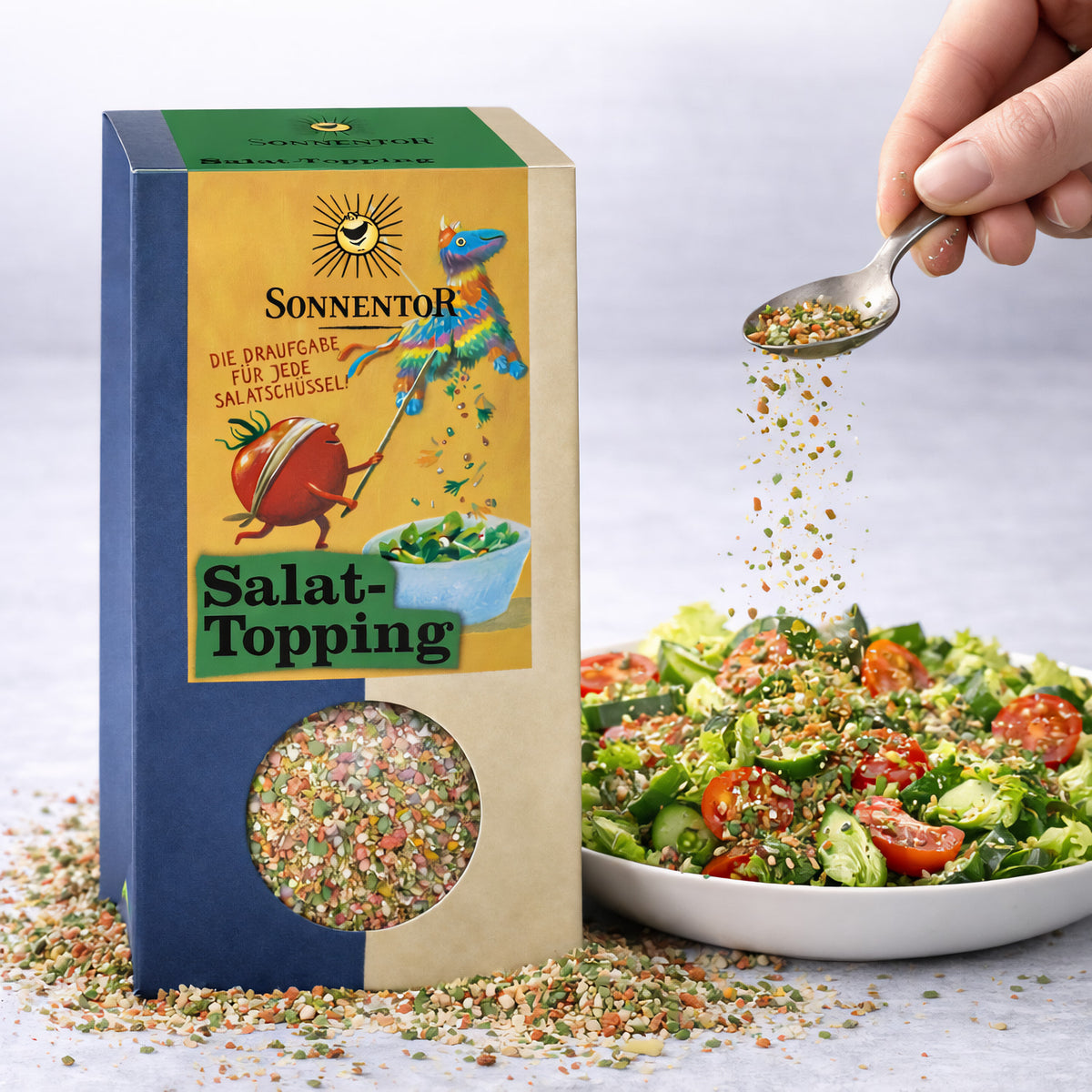 Hand streut Sonnentor Salattopping aus einem Löffel über einen frischen Salat mit Tomaten und Gurken, Packung im Vordergrund.
