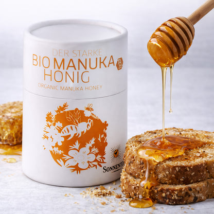 Sonnentor starke Manukahonig 250g neben zwei Brotscheiben, Honig wird mit Honiglöffel als Frühstück-Aufstrich auf das Brot geträufelt.