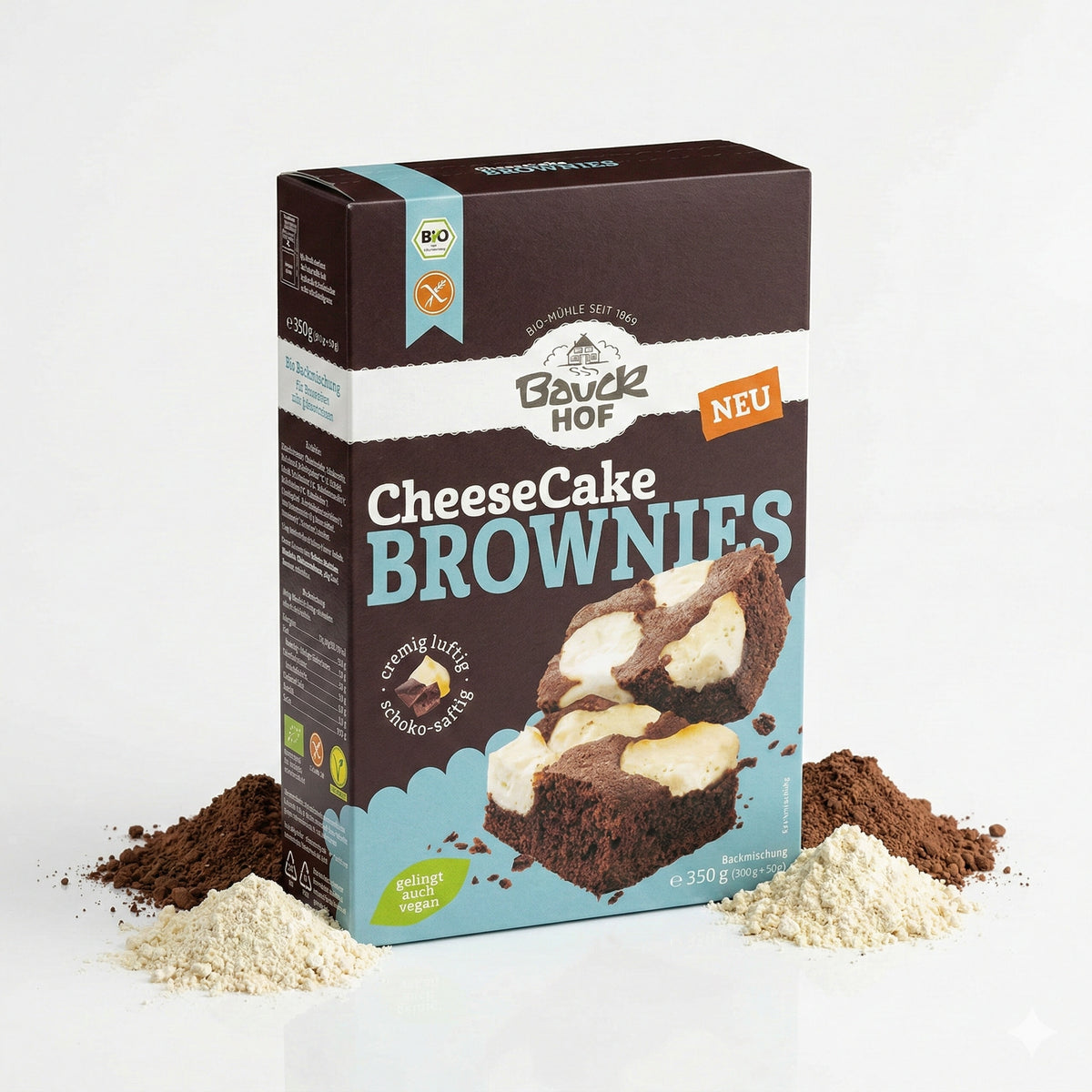 Packung Bauckhof Cheesecake-Brownies Backmischung glutenfrei 350g, stehend vor hellgrauem Hintergrund, umgeben von Kakaopulver und Mehlhäufchen.