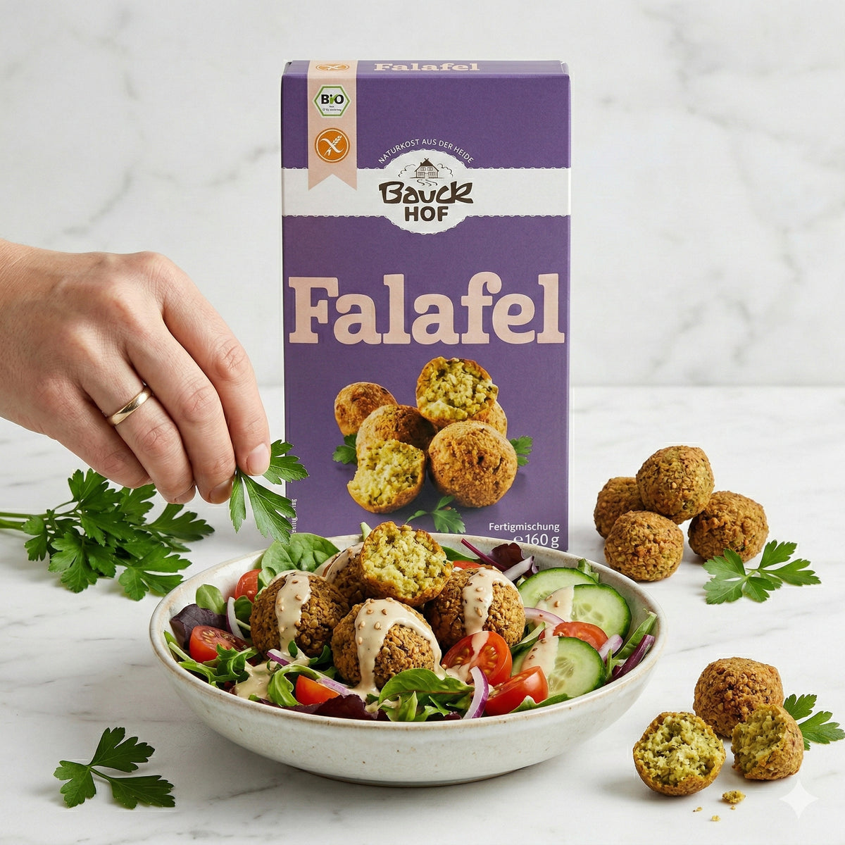 Bauckhof Falafel Mischung glutenfrei hinter einem Salatteller mit Falafel, Tomaten, Gurken und einer Hand, die Petersilie darüber streut.