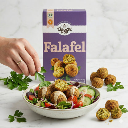 Bauckhof Falafel Mischung glutenfrei hinter einem Salatteller mit Falafel, Tomaten, Gurken und einer Hand, die Petersilie darüber streut.