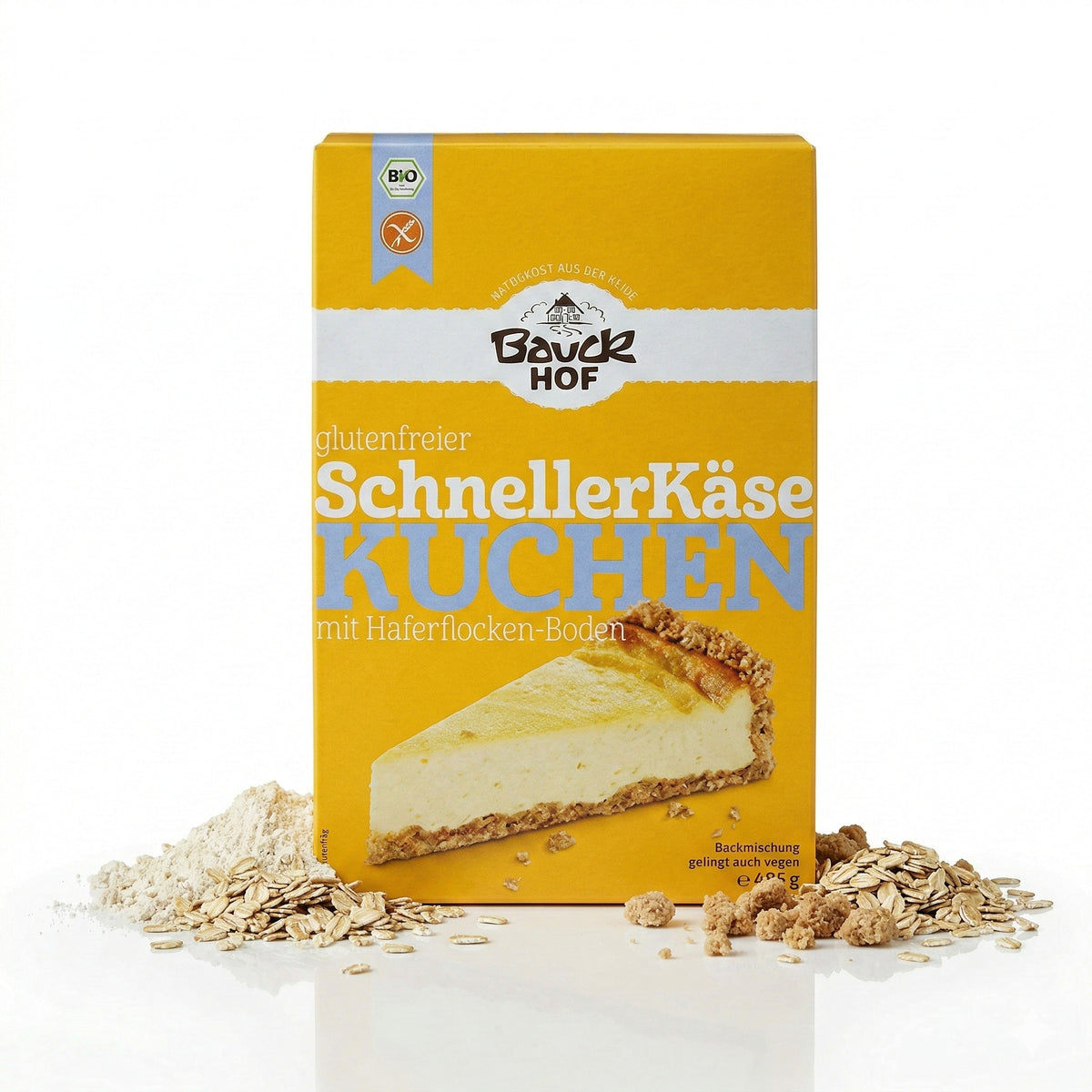 Packung Bauckhof schnelle Käsekuchen Backmischung glutenfrei 485g, stehend vor weißem Hintergrund, daneben Haferflocken und Mehlhäufchen.