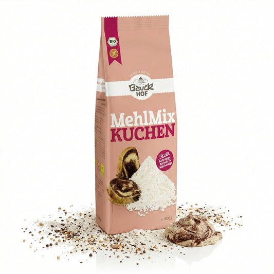 Bauckhof Mehl Mix Kuchen glutenfrei 800g, stehende Packung auf weißem Hintergrund, umgeben von Mehl, Körnern und einem Klecks Teig, vorne Produktabbildung mit Kuchenstücken.
