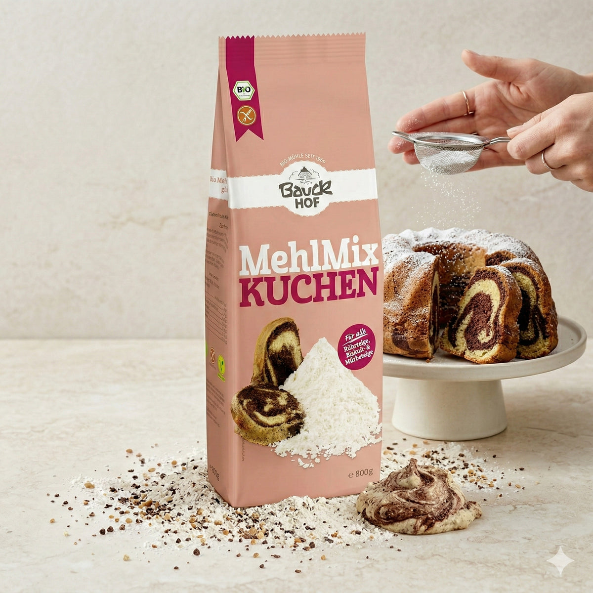 Bauckhof Mehl Mix Kuchen glutenfrei 800g, Packung auf Tisch, im Hintergrund Marmorkuchen auf Tortenplatte, Hände sieben Puderzucker über den Kuchen, vorne Mehl und Teig.