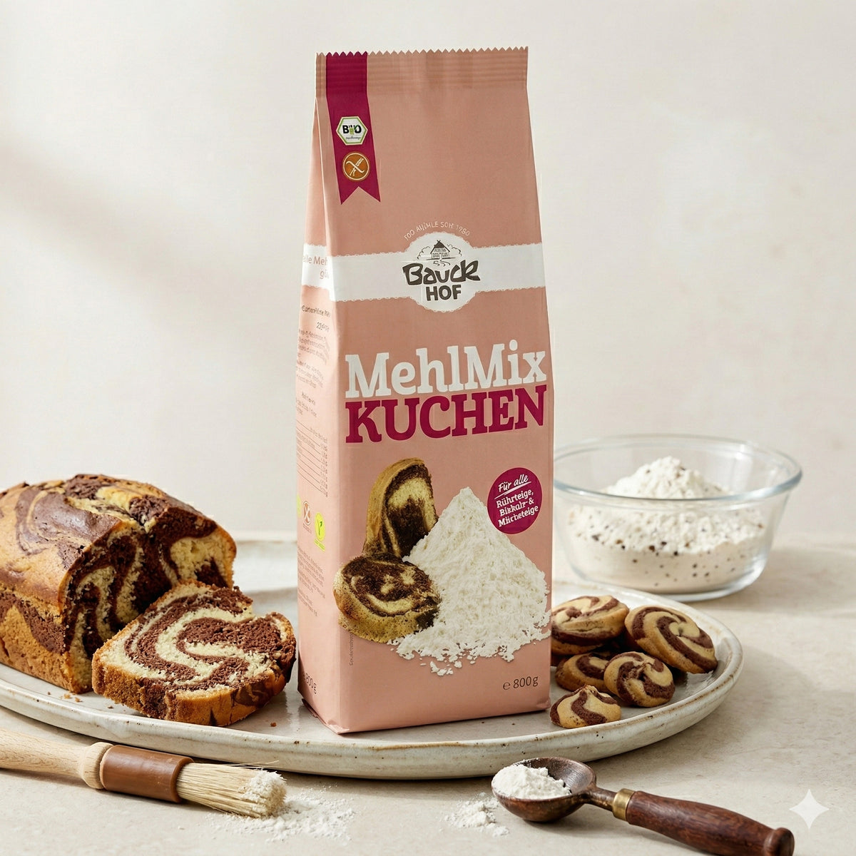 Bauckhof Mehl Mix Kuchen glutenfrei 800g, Packung auf Tablett mit angeschnittenem Marmorkuchen, Schüssel mit Mehl und einige Kekse, daneben Backpinsel und Holzlöffel mit Mehl.