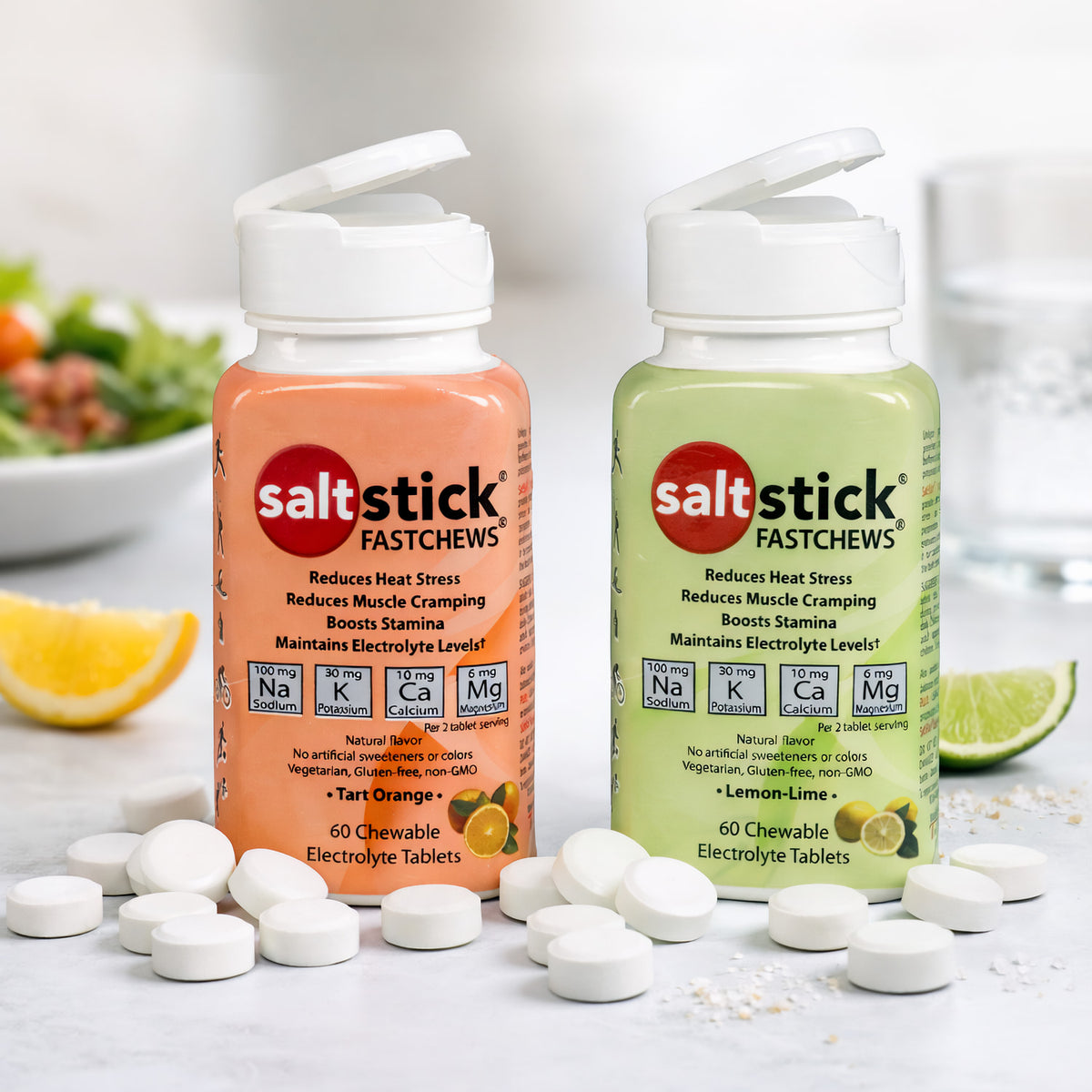 SaltStick Fast Chews Elektrolyt-Kautabletten in Orange- und Zitronen-Limetten-Geschmack, im Vordergrund weiße Kautabletten, im Hintergrund ein Salatteller und ein Glas Wasser.