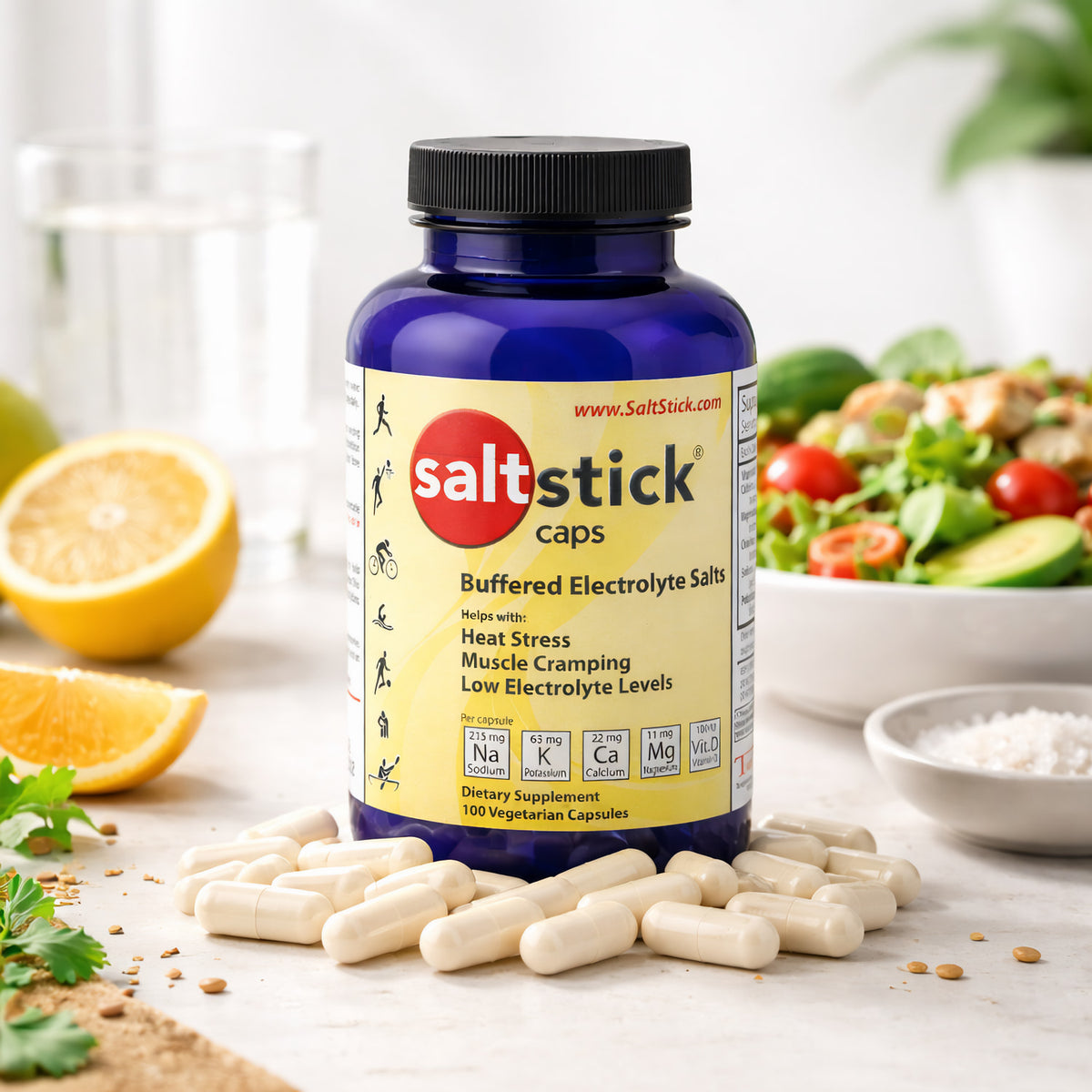 SaltStick Kapseln vor einer blauen Dose, im Hintergrund Zitronenhälften, ein Glas Wasser und ein Salat, Kapseln liegen auf dem Tisch.