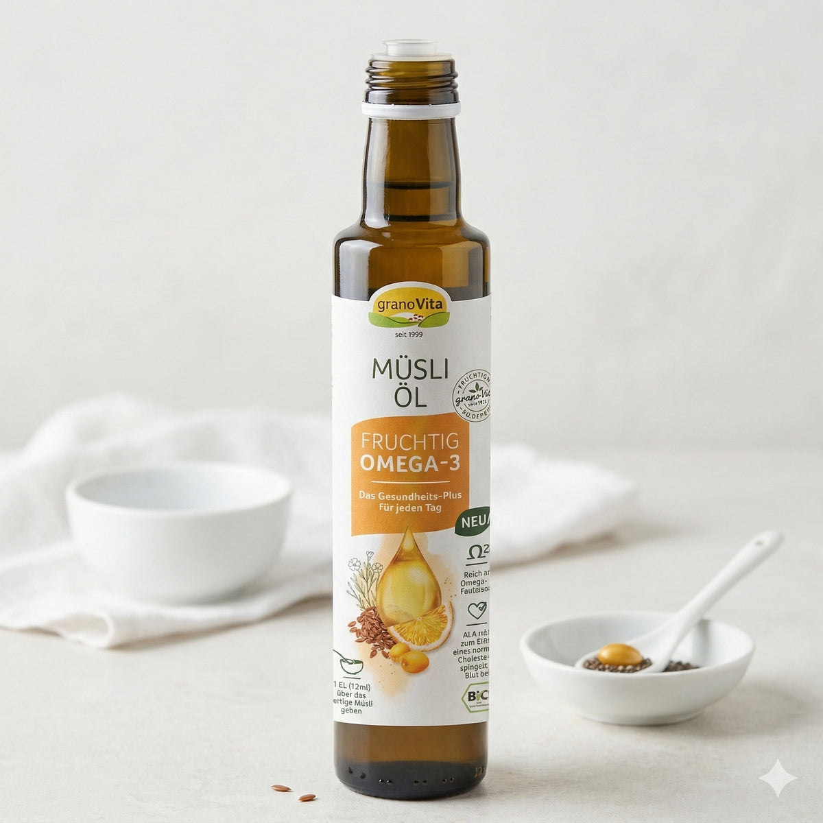 geöffnete Flasche grano vita müsli öl fruchtig auf hellem Untergrund, im Hintergrund weiße Schale mit Löffel und etwas Öl, daneben Leinsamen