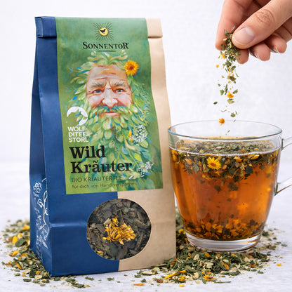 Bio Wildkräuter-Tee von Sonnentor in einer Packung, frische Kräuter für aromatischen Genuss, geöffnete Teetasse mit Kräutern.