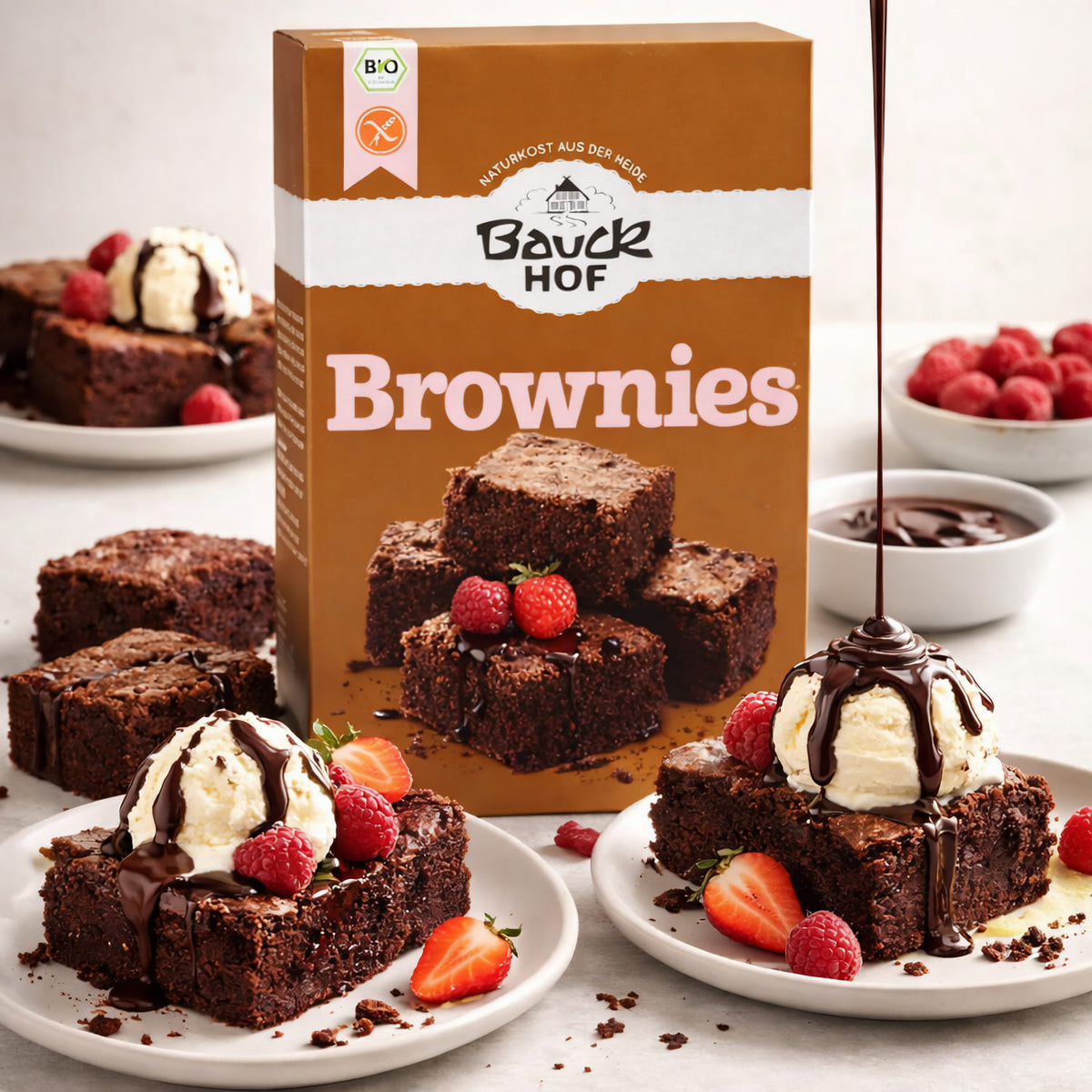 Bauckhof Brownies glutenfrei serviert auf Tellern mit Vanilleeis, Schokosauce, Erdbeeren und Himbeeren, Packung im Hintergrund.