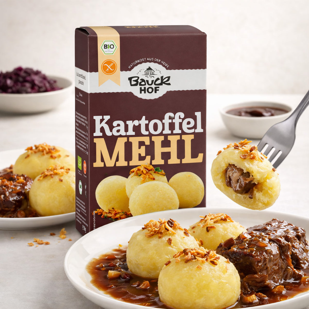 Bauckhof kartoffelmehl Packung neben einem Teller mit Kartoffelklößen, Bratensoße und Fleisch, ein Kloß wird mit einer Gabel aufgespießt, gefüllt mit Soße.