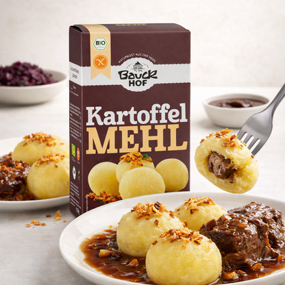 Bauckhof kartoffelmehl Packung neben einem Teller mit Kartoffelklößen, Bratensoße und Fleisch, ein Kloß wird mit einer Gabel aufgespießt, gefüllt mit Soße.