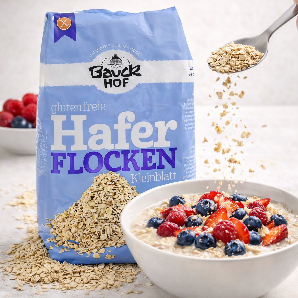 Bauckhof Haferflocken Kleinblatt neben einer Schale Porridge mit Beeren, Haferflocken werden mit einem Löffel hinzugefügt.