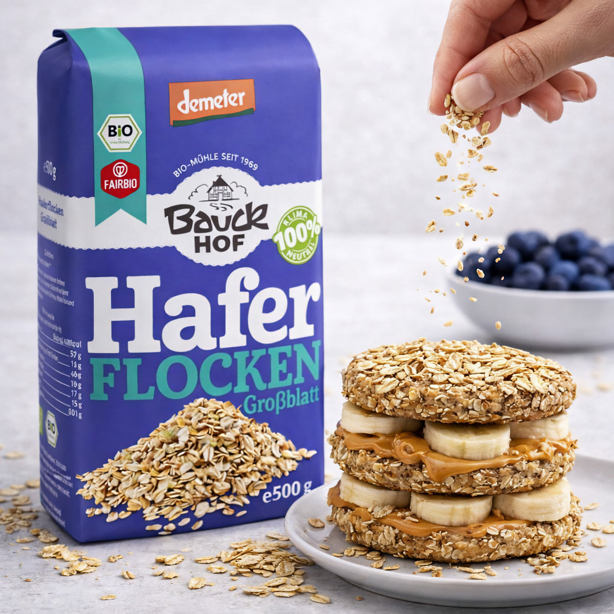 Bauckhof Haferflocken Großblatt Demeter 500g neben einem Teller mit Haferflocken-Sandwich aus Banane und Erdnussmus, Hand streut Haferflocken darüber.