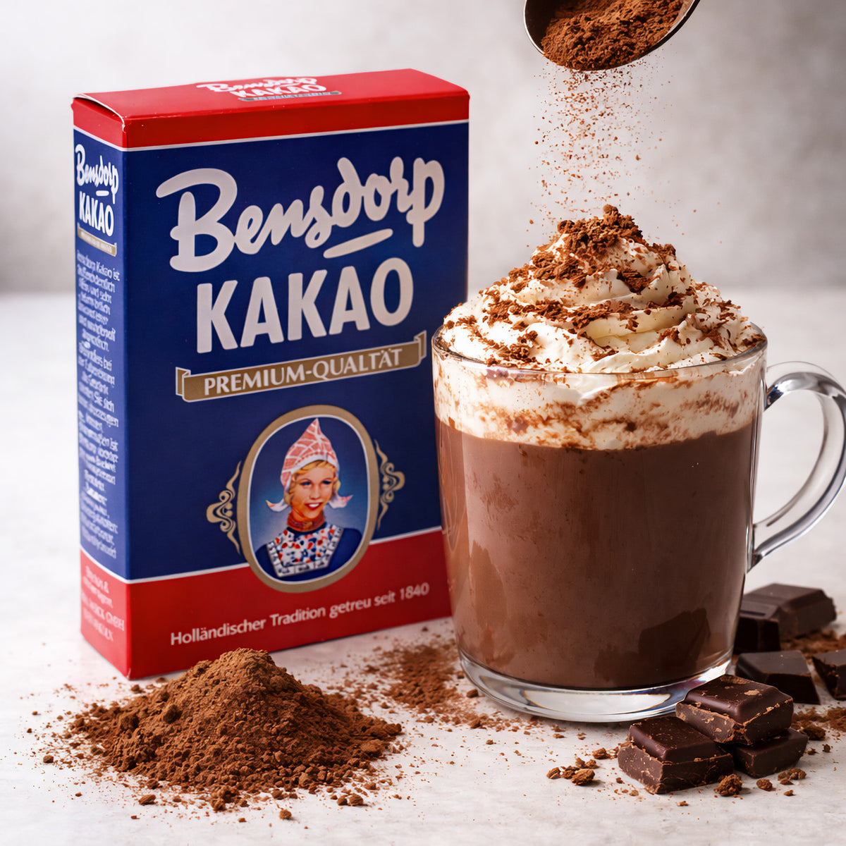 Bensdorp Kakao Getränkepulver neben einer Tasse Trinkschokolade mit Sahne, Kakaopulver und Schokoladenstücken.