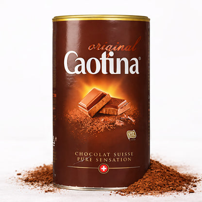 Eine Dose Caotina Original Trinkschokolade 500g steht auf einer Unterlage aus verstreutem Kakaopulver, das Etikett mit Schokoladenstücken ist gut sichtbar.