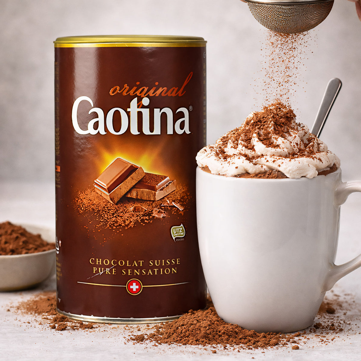 Caotina Original Getränkepulver neben einer Tasse heißer Schokolade mit Sahne, bestreut mit Kakaopulver, im Vordergrund Kakaopulver auf dem Tisch.
