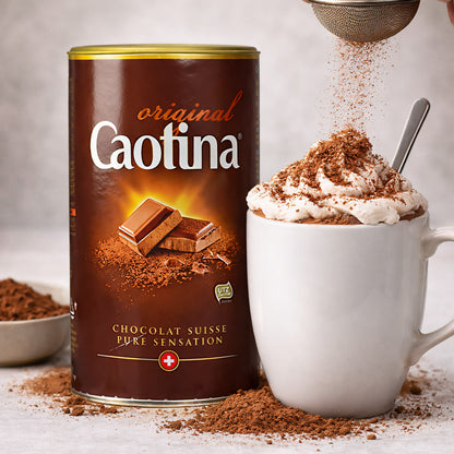 Caotina Original Getränkepulver neben einer Tasse heißer Schokolade mit Sahne, bestreut mit Kakaopulver, im Vordergrund Kakaopulver auf dem Tisch.