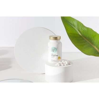 cosphera biotin tabletten dose auf weißem podest, einige tabletten davor, pflanzenblatt im hintergrund