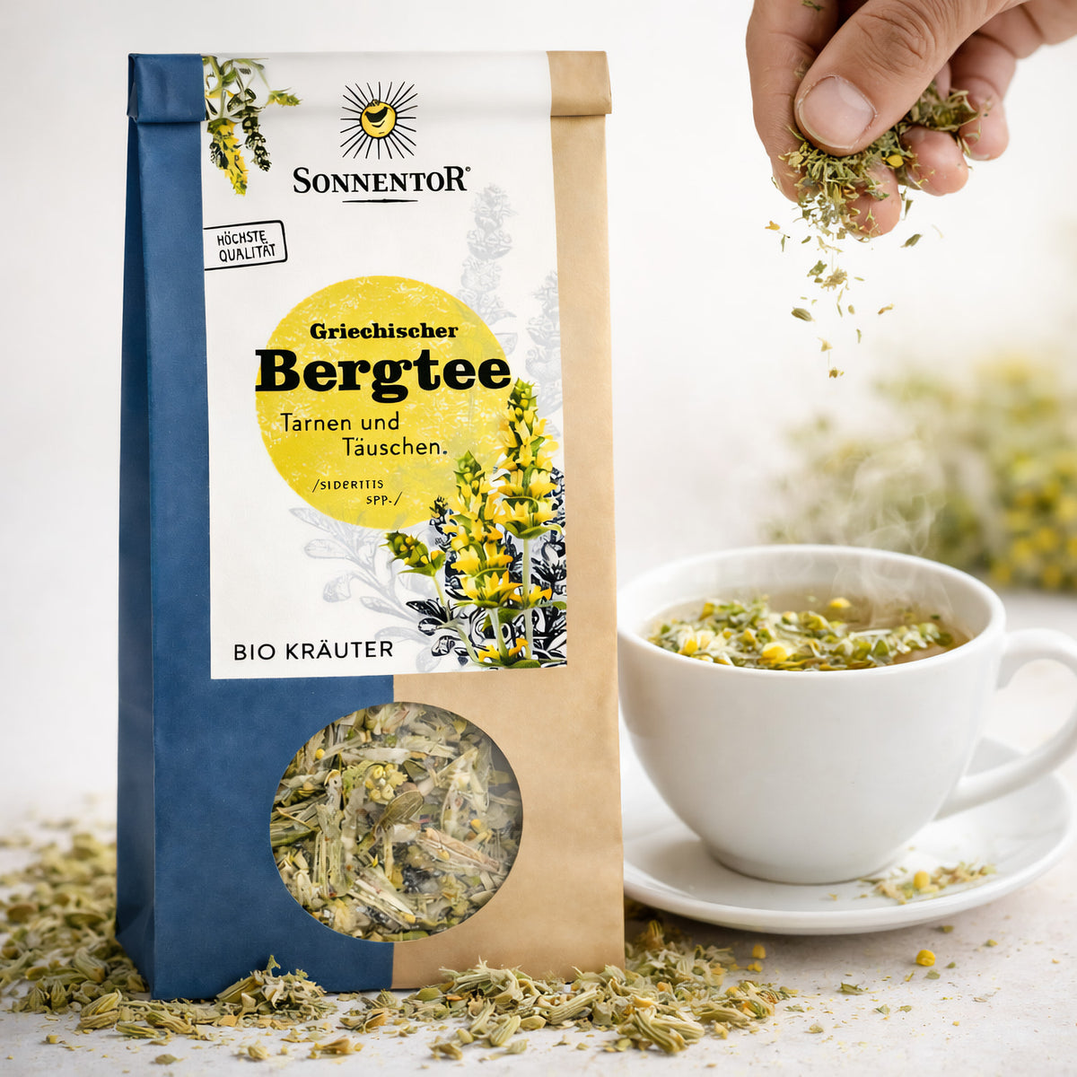 Sonnentor griechischer Bergtee lose neben einer dampfenden Tasse, Hand streut Kräutertee in die Tasse, Verpackung und Kräuter sichtbar.