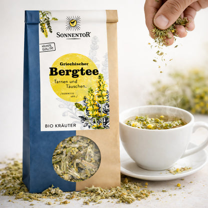 Sonnentor griechischer Bergtee lose neben einer dampfenden Tasse, Hand streut Kräutertee in die Tasse, Verpackung und Kräuter sichtbar.