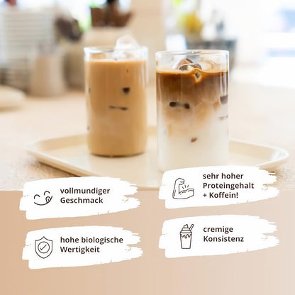 Zwei Gläser mit eisgekühltem proteinkaffee auf einem Tablett, links helles, rechts geschichtetes Kaffeegetränk, im Hintergrund verschwommene Küche.