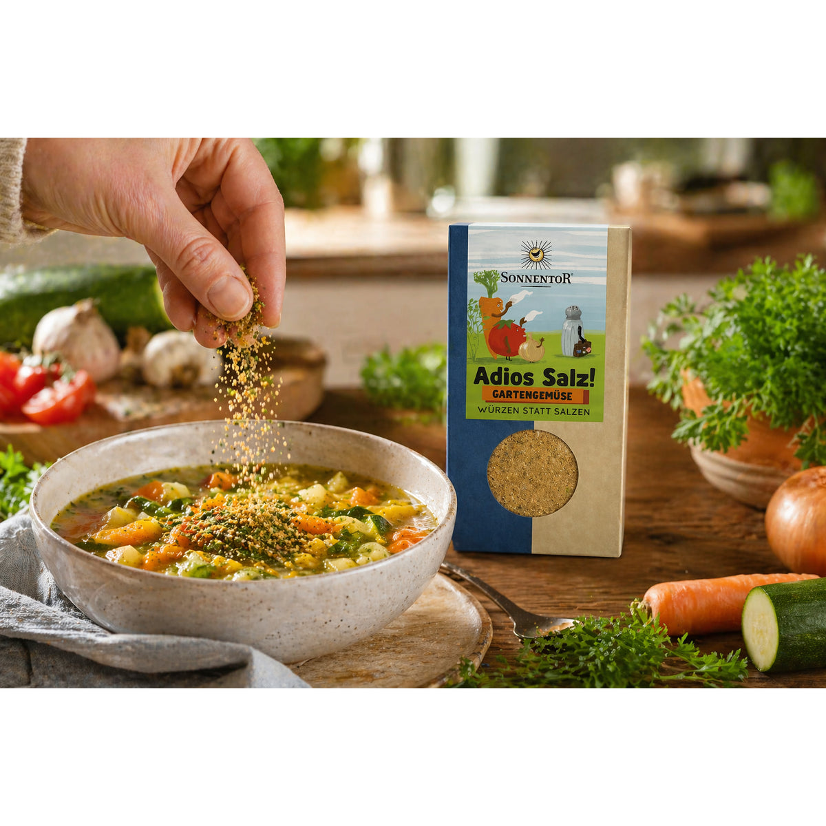 Hand streut Sonnentor Adios Salz! Gartengemüse Gewürz über einen Teller Gemüsesuppe, Verpackung steht daneben auf dem Tisch.