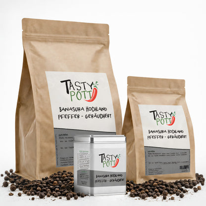 Drei Tasty Pott Banasura Hochlandpfeffer geräuchert Verpackungen im Vordergrund, zwei braune Nachfüllbeutel und eine silberne Dose, umgeben von schwarzen Pfefferkörnern.