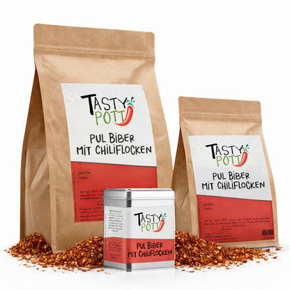 Drei Packungen Tasty Pott Pul Biber mit Chiliflocken in unterschiedlichen Größen, stehend auf einem Haufen Chiliflocken.