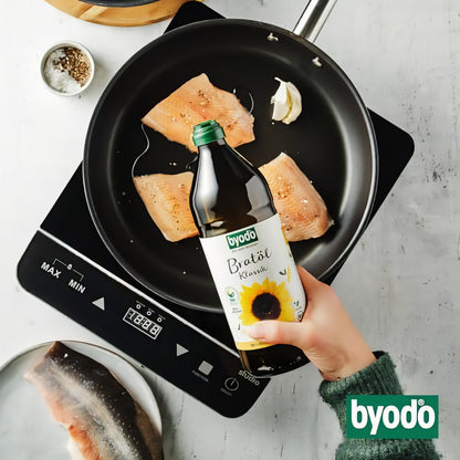 Hand gießt Byodo Frittieröl aus High-Oleic Sonnenblumenkernen in eine Pfanne mit Fischfilets auf einem Herd.