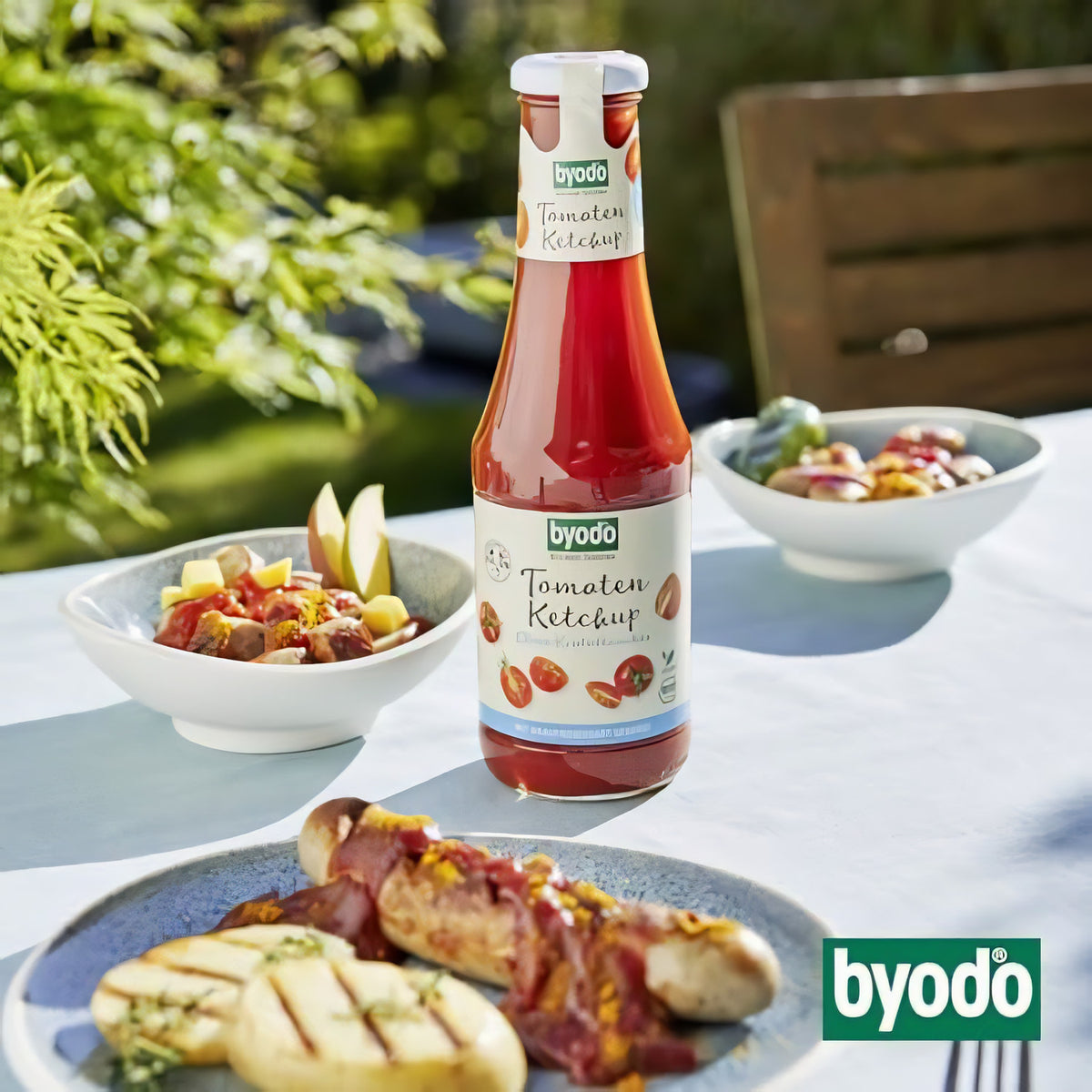 Byodo Tomaten Ketchup Flasche auf gedecktem Tisch im Garten, umgeben von Grillgericht und Salatschalen.