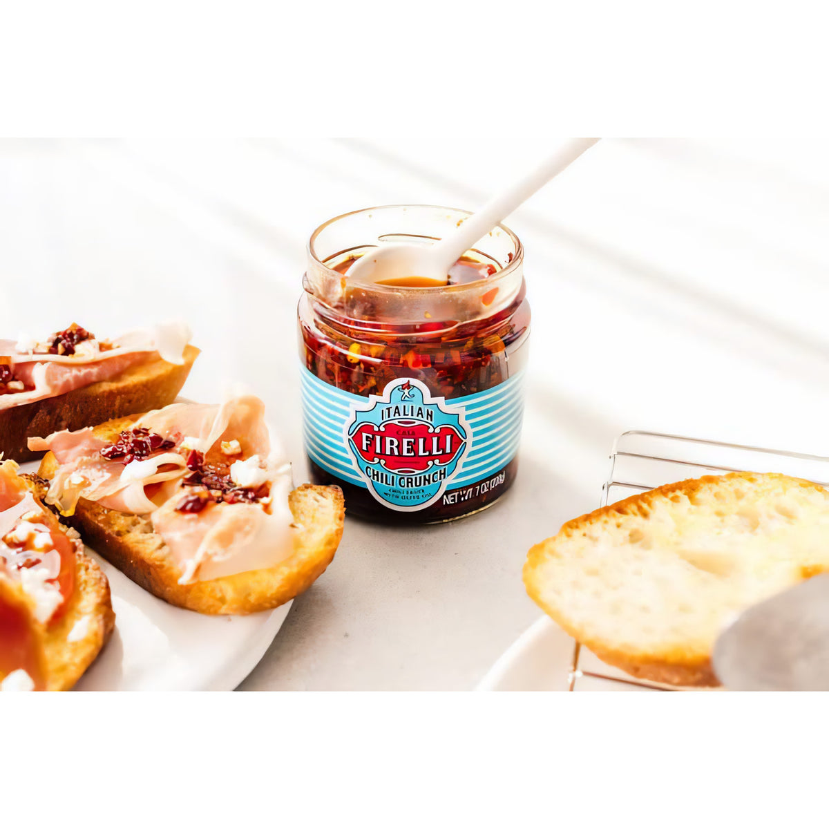 Geöffnetes Glas Casa Firelli Chili Crunch mit Löffel, daneben belegte Brotscheiben als Topping-Idee.