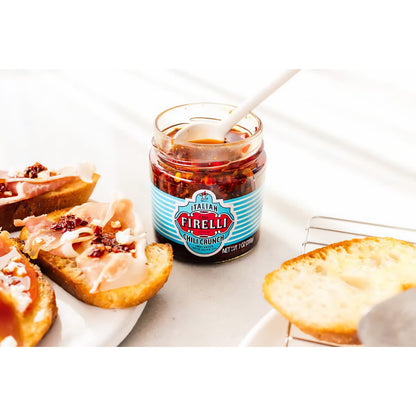 Geöffnetes Glas Casa Firelli Chili Crunch mit Löffel, daneben belegte Brotscheiben als Topping-Idee.