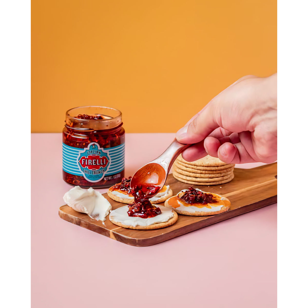 Casa Firelli Chili Crunch Glas auf Holzbrett, ein Löffel Chili Crunch wird auf Cracker mit Frischkäse gegeben.