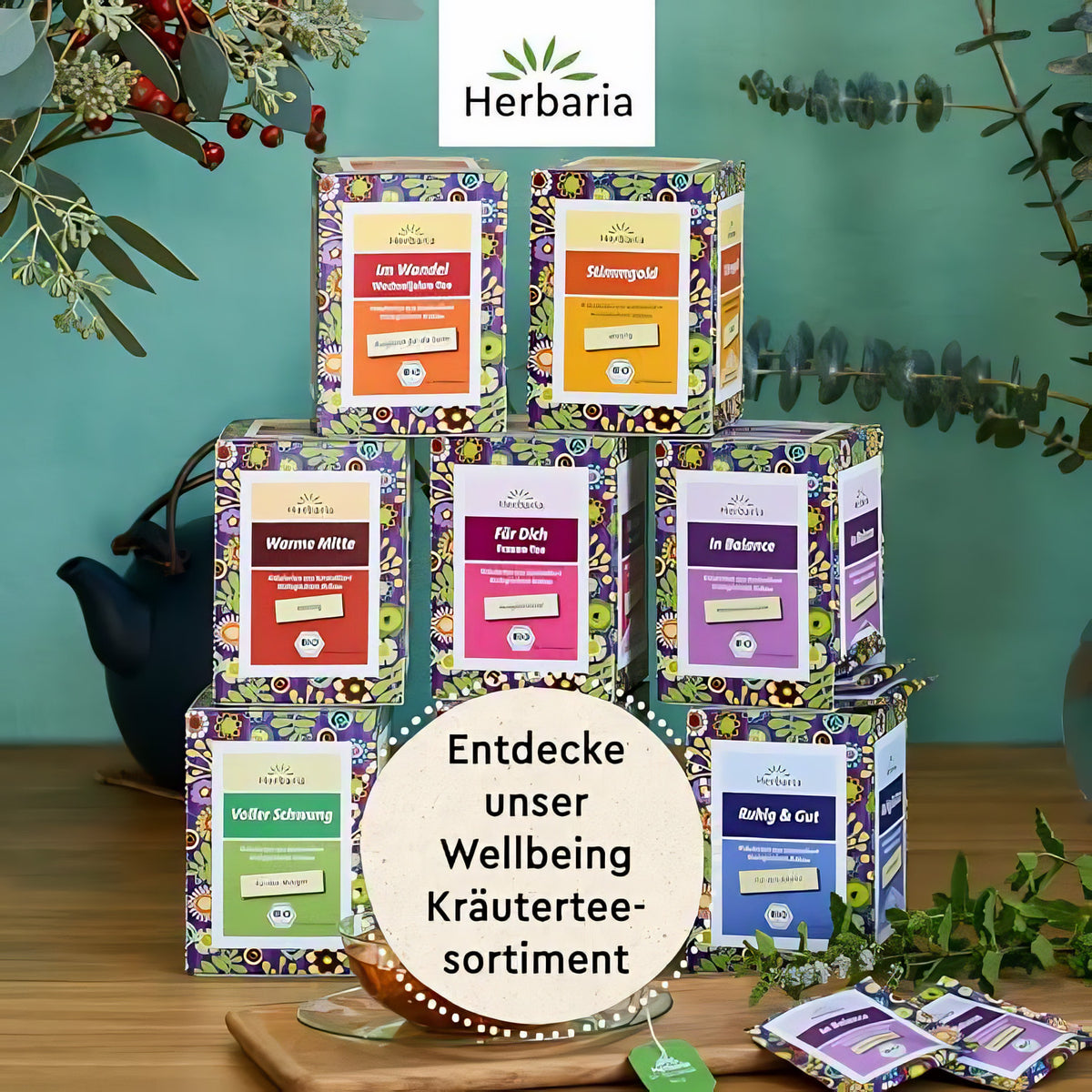 Verschiedene Herbaria Kräutertee-Packungen, darunter Im Wandel 15FB, als Pyramide auf einem Holztisch vor grünem Hintergrund arrangiert.
