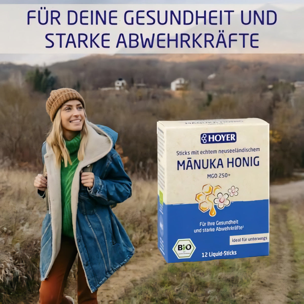 Frau im Outdoor-Outfit neben einer großen Packung HOYER Manuka Honig Liquid-Sticks vor herbstlicher Landschaft.