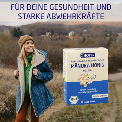 Frau im Outdoor-Outfit neben einer großen Packung HOYER Manuka Honig Liquid-Sticks vor herbstlicher Landschaft.