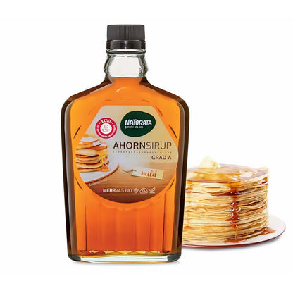 Naturata Ahornsirup Grad A in Glasflasche, vorne stehend, daneben ein Stapel Pancakes mit Sirup auf weißem Teller.