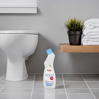 Klar WC-Reiniger 750ml ohne Duft steht auf hellgrauen Badezimmerfliesen neben einer Toilette, Regal mit Handtüchern und Pflanze im Hintergrund.
