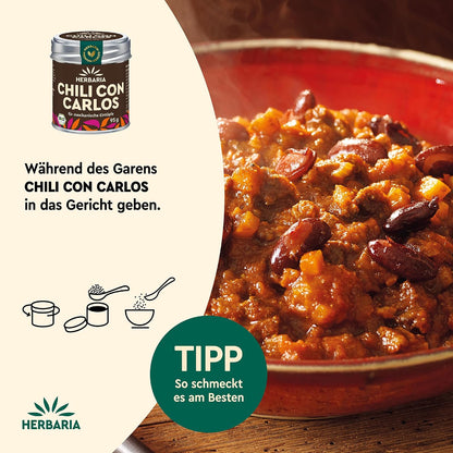 Nahaufnahme eines dampfenden Chili-Eintopfs in einem roten Topf, links daneben Herbaria Chili con Carlos Gewürzdose und Anwendungshinweis.