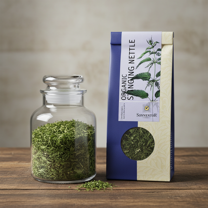 Capture a high-resolution close-up of Sonnentor Bio Brennnessel Tee lose in a clear, elegant glass jar, illuminated by softbox lighting that highlights the vibrant green of the tea leaves. The scene is set against a minimalist, neutral-colored background w