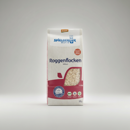 A professional commercial studio shot emphasizing the eco-conscious packaging of Spielberger Bio Roggenflocken (500g). The package is set against a sleek, minimalist background with a light source creating rim lighting that highlights its contours and natu