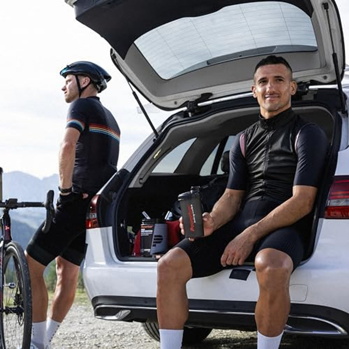 Zwei Radfahrer in Sportkleidung, einer sitzt am offenen Kofferraum eines Autos mit Powerbar Build Whey Protein Black Line Shaker und Dose Molkenproteine.